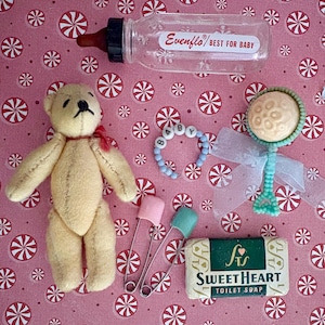 Könnte beinhalten: Eine Sammlung von Babyartikeln auf rosa Hintergrund mit Zuckerstangenmuster. Enthält einen Teddybär, eine Babyflasche, eine Rassel, ein Armband mit der Aufschrift "BABY", Sicherheitsnadeln und ein Stück "Sweet Heart" Seife.