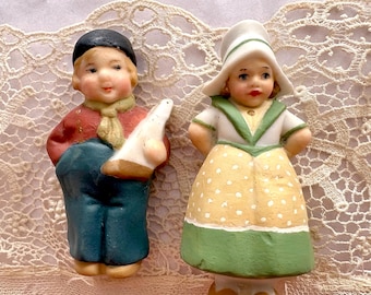 Pode incluir: Duas figuras de cerâmica de crianças em trajes tradicionais holandeses. Uma figura usa um boné preto, um casaco vermelho e calças azuis, segurando um objeto branco. A outra usa um chapéu branco, uma blusa branca com acabamento verde e um vestido amarelo com bolinhas.