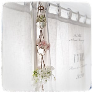 Peut inclure: Un porte-plante en macramé avec trois globes en verre, contenant des fleurs roses et blanches. Le porte-plante est suspendu devant un rideau blanc transparent.