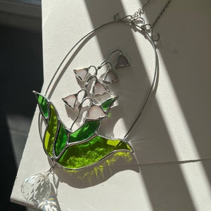 Puede incluir: Un atrapasol de vidrio tintado con un diseño de lirio de los valles y un prisma de cristal colgando de la parte inferior. El lirio de los valles está hecho de vidrio verde y blanco y está rodeado por un marco de metal plateado.