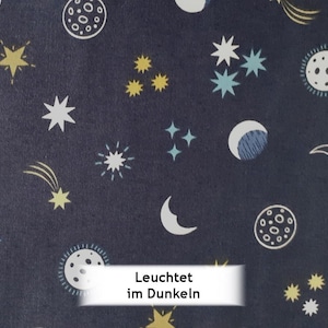 Könnte beinhalten: Dunkelblauer Stoff mit einem sich wiederholenden Muster von Himmelskörpern. Das Design umfasst weiße, gelbe und hellblaue Sterne, Monde und Planeten. Der Text "Leuchtet im Dunkeln" wird auf einem weißen Banner angezeigt.