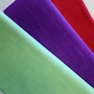 Könnte beinhalten: Nahaufnahme von drei Stoffmustern in Grün, Lila und Rot. Die Stoffe sind diagonal angeordnet und zeigen ihre Texturen und Farben. Der grüne Stoff befindet sich unten, gefolgt von Lila und Rot.