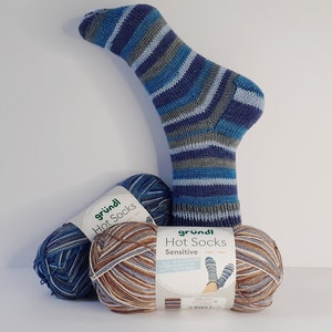 Könnte beinhalten: Eine gestreifte Socke in Blau-, Grau- und Weißtönen, mit zwei Knäueln Garn in passenden Farben. Die Garnetiketten tragen die Aufschrift "Gründl Hot Socks Sensitive".