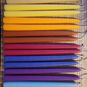 May include: A set of 14 taper candles in various colors, including white, beige, yellow, orange, brown, red, purple, blue, and green. The candles are arranged in a rainbow pattern.
