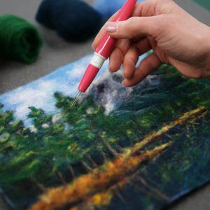 May include: A person uses a needle tool to create a felt landscape art piece. The artwork depicts a forest scene with green trees, a blue sky, and a brown river.