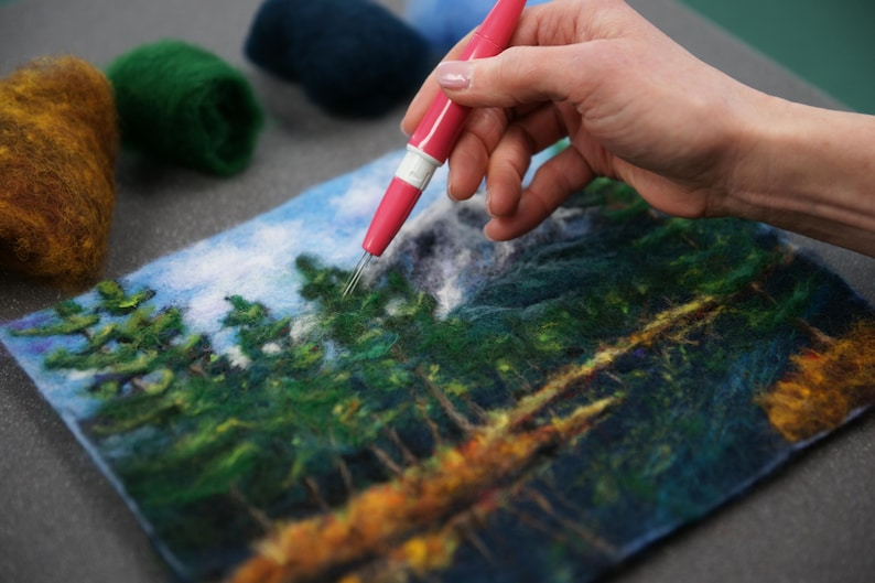 May include: A person uses a needle tool to create a felt landscape art piece. The artwork depicts a forest scene with green trees, a blue sky, and a brown river.