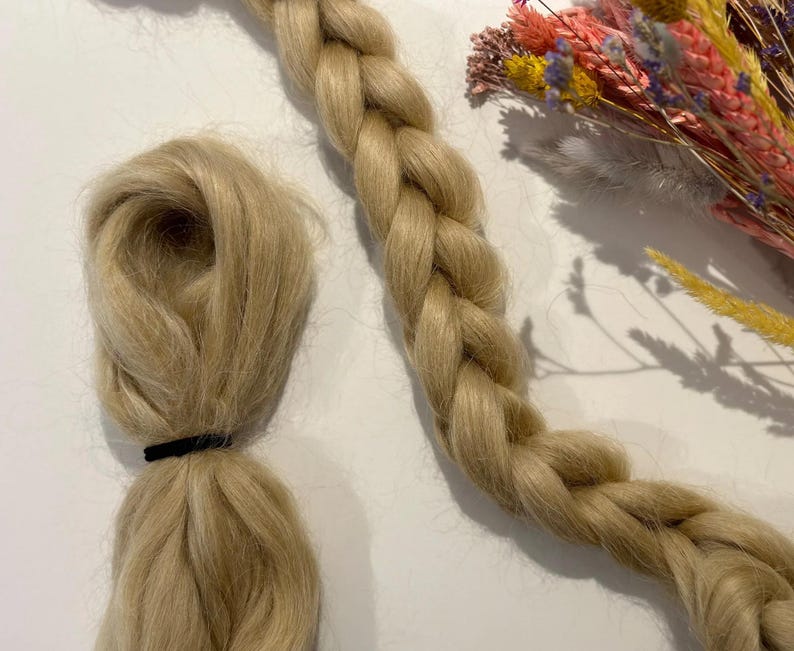 K&ouml;nnte beinhalten: Zwei Str&auml;hnen hellblondes Haar, eine geflochten und die andere locker, werden vor einem wei&szlig;en Hintergrund gezeigt. Die lockere Str&auml;hne ist mit einem schwarzen Gummiband gebunden.