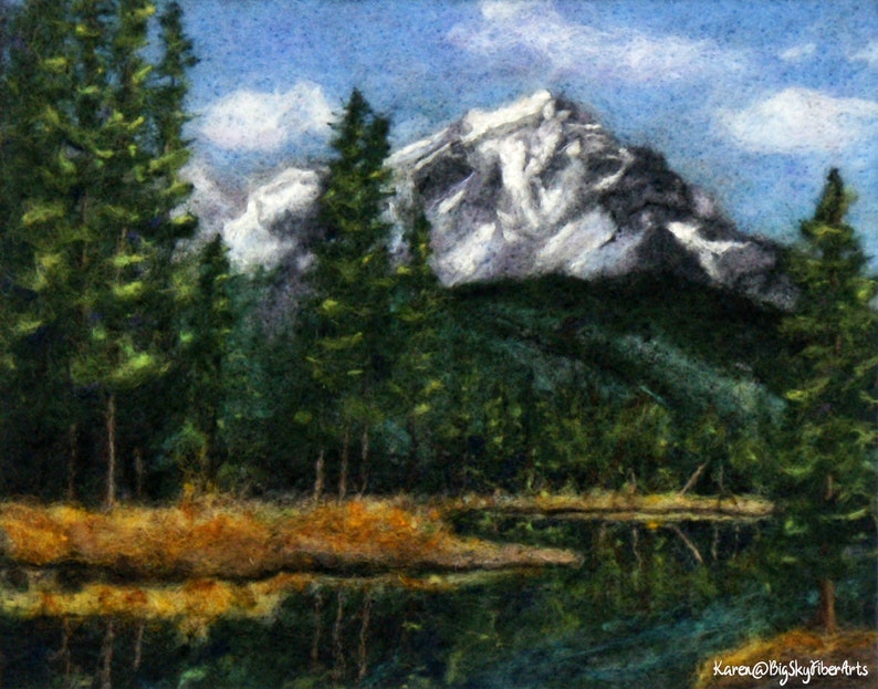May include: A needle felted artwork depicting a mountain range with snow-capped peaks, a lake reflecting the mountains, and a foreground of evergreen trees.