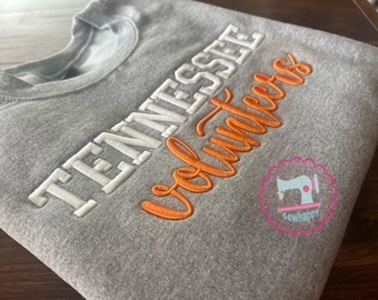 May include: A gray sweatshirt with the words "TENNESSEE Volunteers" embroidered in white and orange. A pink and blue logo is in the lower right corner. The sweatshirt is folded.