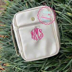 Puede incluir: Un bolso bandolera cuadrado dorado con cierre de cremallera y asa. El bolso presenta un monograma bordado en rosa y un bot&oacute;n dorado. El bolso est&aacute; sobre hierba verde.