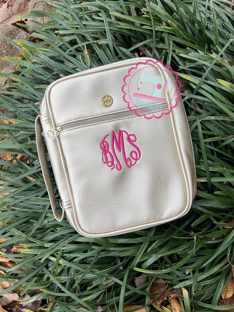 Puede incluir: Un bolso bandolera cuadrado dorado con cierre de cremallera y asa. El bolso presenta un monograma bordado en rosa y un bot&oacute;n dorado. El bolso est&aacute; sobre hierba verde.