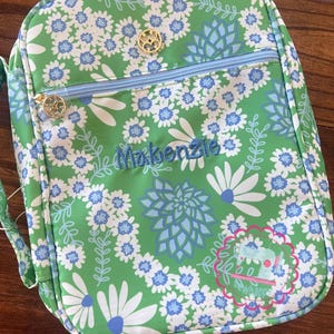 Puede incluir: Bolsa de almuerzo con estampado floral verde con flores blancas y azules. La bolsa tiene una cremallera azul claro y tiradores dorados. El nombre "Makenzie" est&aacute; bordado en azul en la parte delantera.