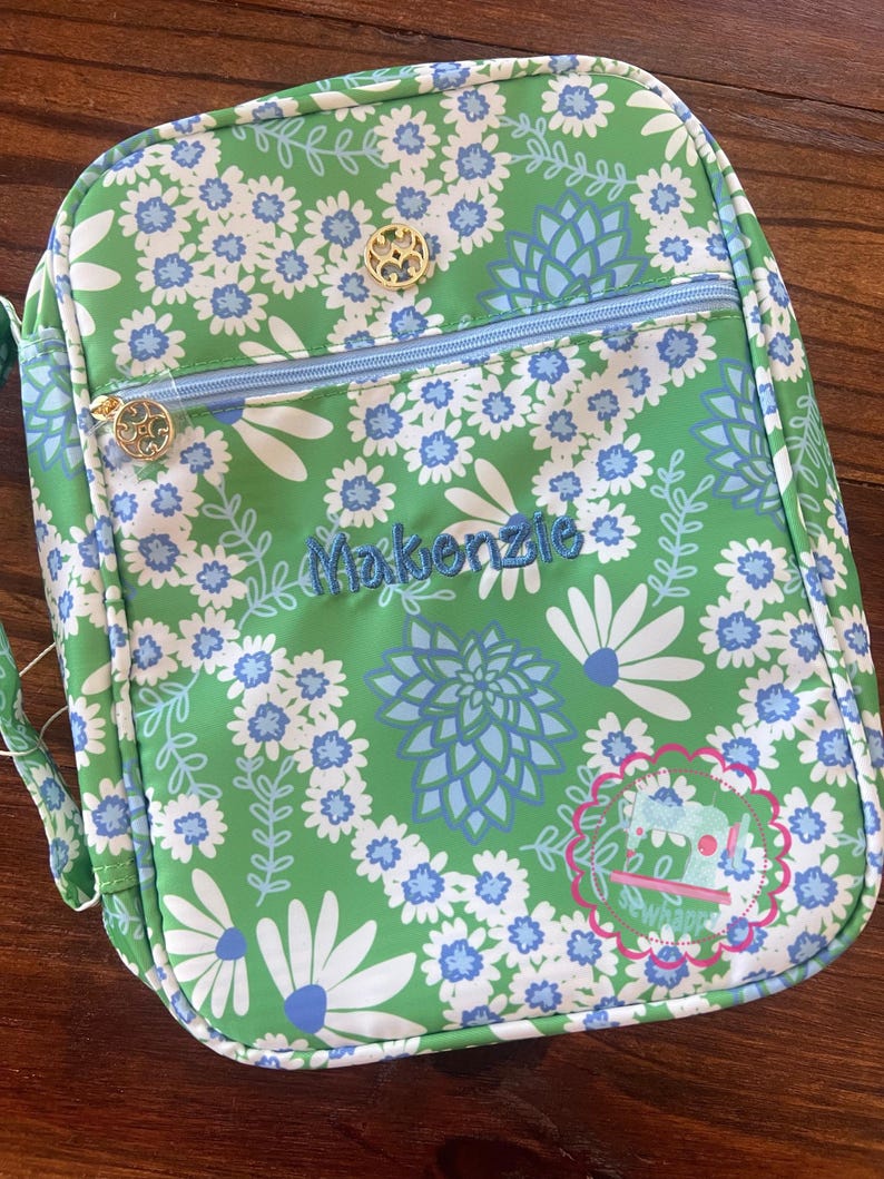 Puede incluir: Bolsa de almuerzo con estampado floral verde con flores blancas y azules. La bolsa tiene una cremallera azul claro y tiradores dorados. El nombre "Makenzie" est&aacute; bordado en azul en la parte delantera.