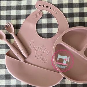 May include: A pink silicone bib with the name "Sophia May" engraved on it. The bib is paired with a pink silicone divided plate and two pink silicone spoons.