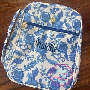 Puede incluir: Una bolsa de almuerzo con estampado floral blanco y azul con cremallera azul y herrajes dorados. El nombre "Natalie" est&aacute; bordado en escritura azul. Un gr&aacute;fico rosa y azul est&aacute; en la esquina inferior derecha. La bolsa tiene un asa y una bandolera.