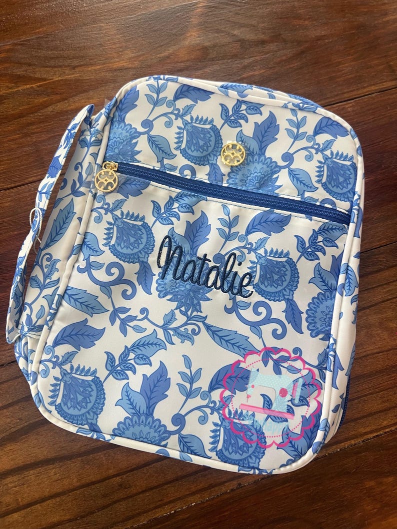 Puede incluir: Una bolsa de almuerzo con estampado floral blanco y azul con cremallera azul y herrajes dorados. El nombre "Natalie" est&aacute; bordado en escritura azul. Un gr&aacute;fico rosa y azul est&aacute; en la esquina inferior derecha. La bolsa tiene un asa y una bandolera.