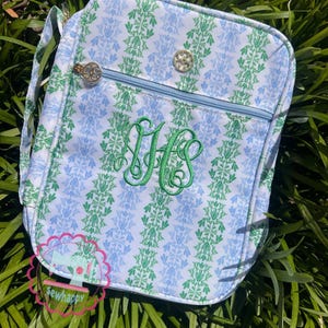 Puede incluir: Un bolso bandolera rectangular con un estampado floral azul y verde. El bolso presenta una cremallera azul claro, herrajes dorados y un monograma verde bordado. El bolso tiene una correa para el hombro y un peque&ntilde;o logotipo que dice "sewhappy".