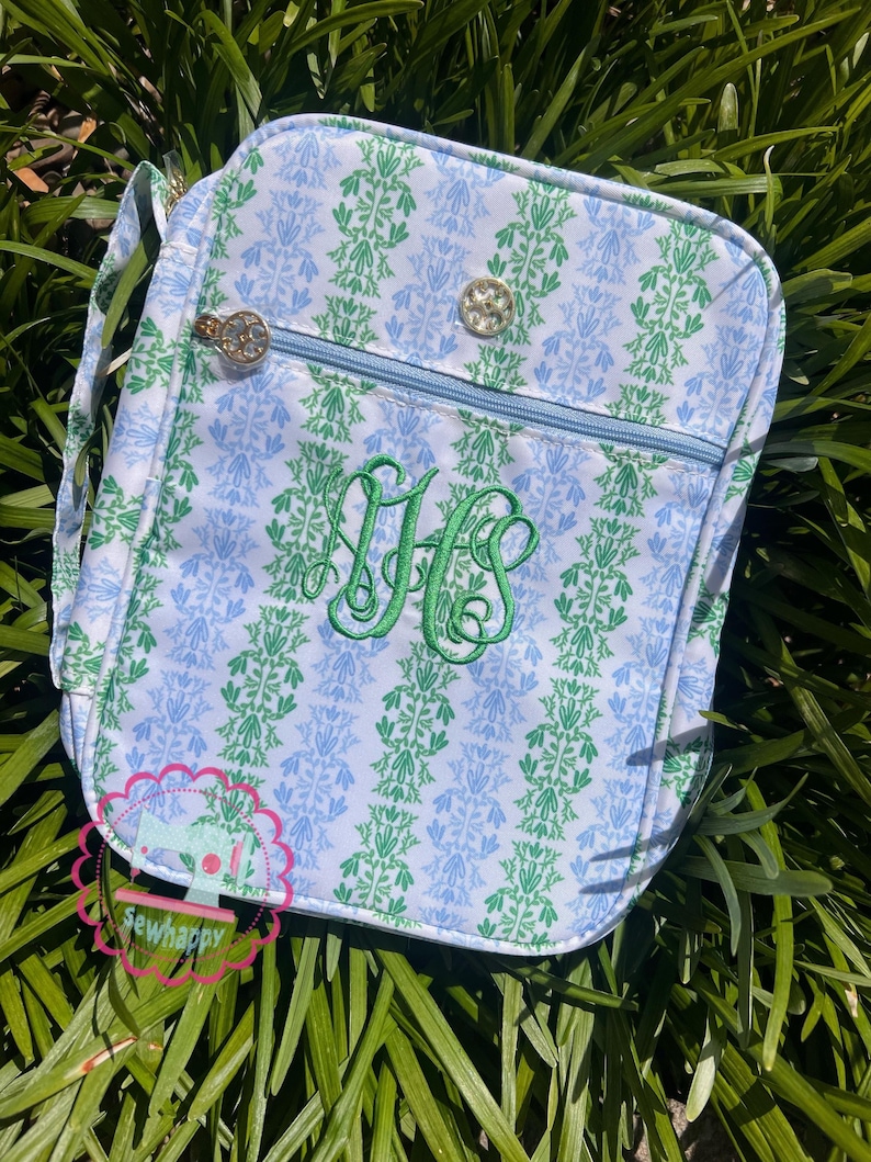 Puede incluir: Un bolso bandolera rectangular con un estampado floral azul y verde. El bolso presenta una cremallera azul claro, herrajes dorados y un monograma verde bordado. El bolso tiene una correa para el hombro y un peque&ntilde;o logotipo que dice "sewhappy".