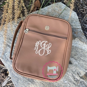 Puede incluir: Un bolso rectangular marr&oacute;n con asa, cremallera y herrajes decorativos dorados. El bolso presenta un monograma blanco y el texto "sewhappy" en un logotipo rosa y blanco.