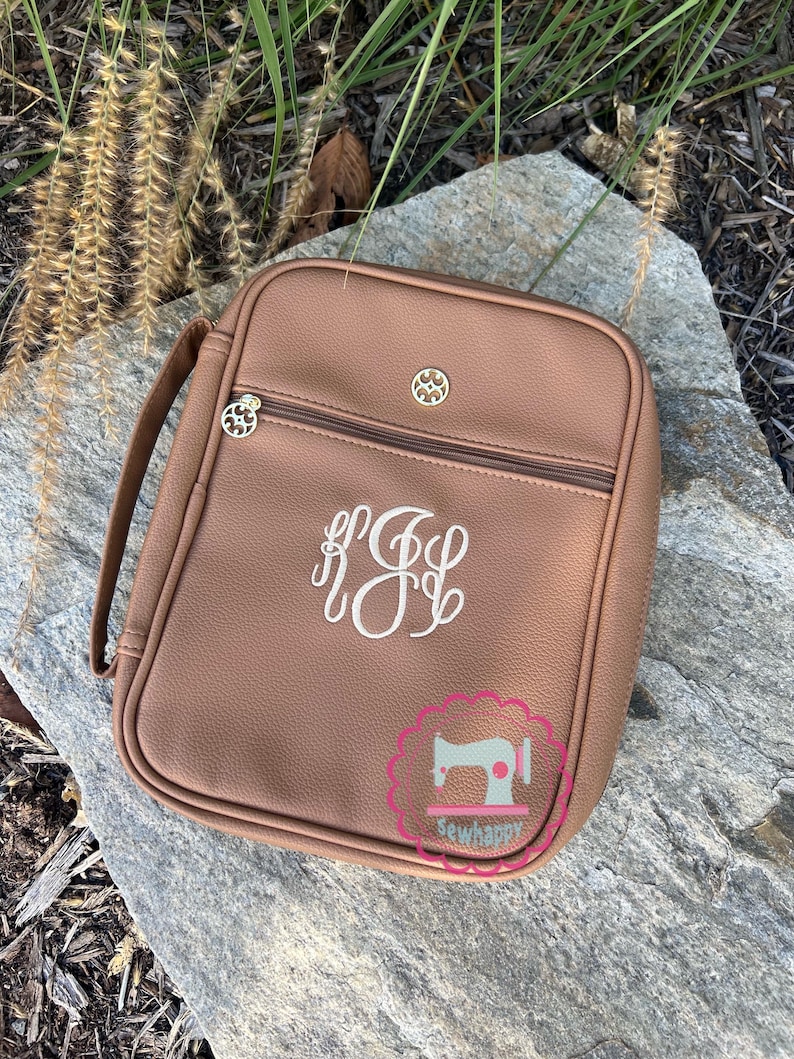 Puede incluir: Un bolso rectangular marr&oacute;n con asa, cremallera y herrajes decorativos dorados. El bolso presenta un monograma blanco y el texto "sewhappy" en un logotipo rosa y blanco.