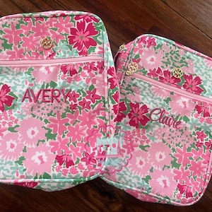 Puede incluir: Dos bolsas de almuerzo con estampado floral con flores rosas, verdes y blancas. Una bolsa tiene el nombre "Avery" bordado en rosa, y la otra "Claire". Ambas bolsas tienen cremalleras y detalles dorados.