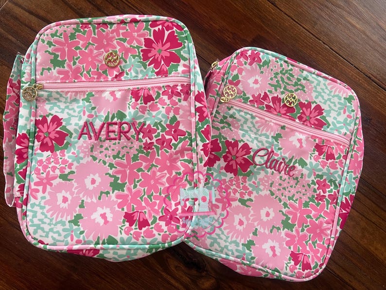 Puede incluir: Dos bolsas de almuerzo con estampado floral con flores rosas, verdes y blancas. Una bolsa tiene el nombre "Avery" bordado en rosa, y la otra "Claire". Ambas bolsas tienen cremalleras y detalles dorados.
