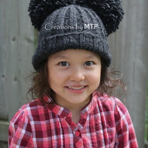 May include: A child wearing a gray knit hat with two large pom-poms and a red and white plaid shirt. The hat has the text "Creations by MTP" on it.