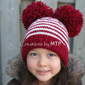 Könnte beinhalten: Ein Kind trägt eine rot-weiß gestreifte Strickmütze mit zwei roten Bommeln. Die Mütze hat den Text "Creations by MTP" am Rand.