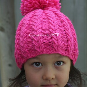 Könnte beinhalten: Eine rosa gestrickte Mütze mit einem Bommel oben. Die Mütze hat ein Zopfmuster und wird von einem Kind mit dunklen Haaren getragen.