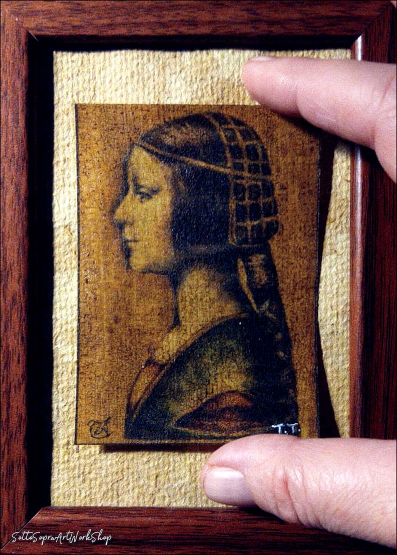 May include: A framed portrait of a woman in profile, rendered in sepia tones. The artwork is set against a textured, light-colored background and framed in dark wood. The image is held by a hand.