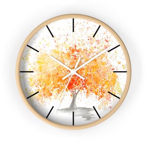 May include: A circular wall clock with a light wood surround. The clock face displays a watercolour painting of a tree with orange and yellow foliage. Black lines mark the hours, and white hands show the time.