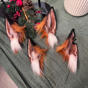 Könnte beinhalten: Vier Fuchsohrenpaare auf silbernen Stirnbändern. Die Ohren sind eine Mischung aus braunem, orangefarbenem und weißem Kunstfell mit schwarzen Spitzen. Die Ohren sind auf einer grünen Stoffoberfläche angeordnet, mit Weihnachtsdekoration im Hintergrund.