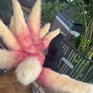 Op de afbeelding: Een harige, meerfingrige hand in cr&egrave;me, roze en perziktinten. Het imitatiebont is lang en pluizig, met een gradi&euml;nteffect. De hand is bevestigd aan een zwarte vorm, wat wijst op een kostuum. De achtergrond toont een stenen terras en groen.