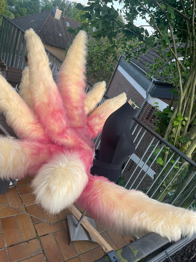 Op de afbeelding: Een harige, meerfingrige hand in cr&egrave;me, roze en perziktinten. Het imitatiebont is lang en pluizig, met een gradi&euml;nteffect. De hand is bevestigd aan een zwarte vorm, wat wijst op een kostuum. De achtergrond toont een stenen terras en groen.