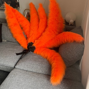 Op de afbeelding: Set van negen feloranje, pluizige vossenstaarten, als kostuumaccessoire. De staarten zijn in een waaier gerangschikt, met een zwarte band voor bevestiging. De imitatiebont is feloranje, geschikt voor cosplay of thema-evenementen.