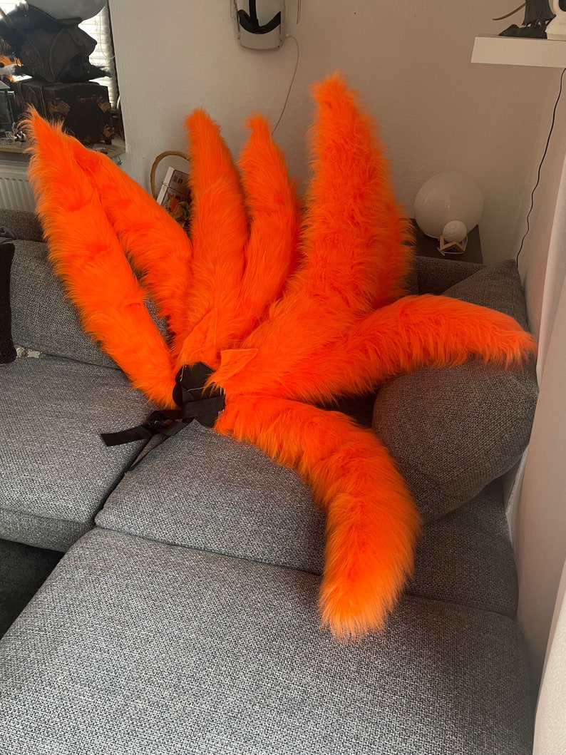 Op de afbeelding: Set van negen feloranje, pluizige vossenstaarten, als kostuumaccessoire. De staarten zijn in een waaier gerangschikt, met een zwarte band voor bevestiging. De imitatiebont is feloranje, geschikt voor cosplay of thema-evenementen.