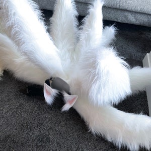 Op de afbeelding: Een pluizige witte vossenstaart met meerdere secties, gecombineerd met bijpassende witte en roze kattenoren en een zwarte hoofdband. De imitatiebont is lang en getextureerd, wat een grillige esthetiek cre&euml;ert. Perfect voor cosplay of kostuumfeesten.