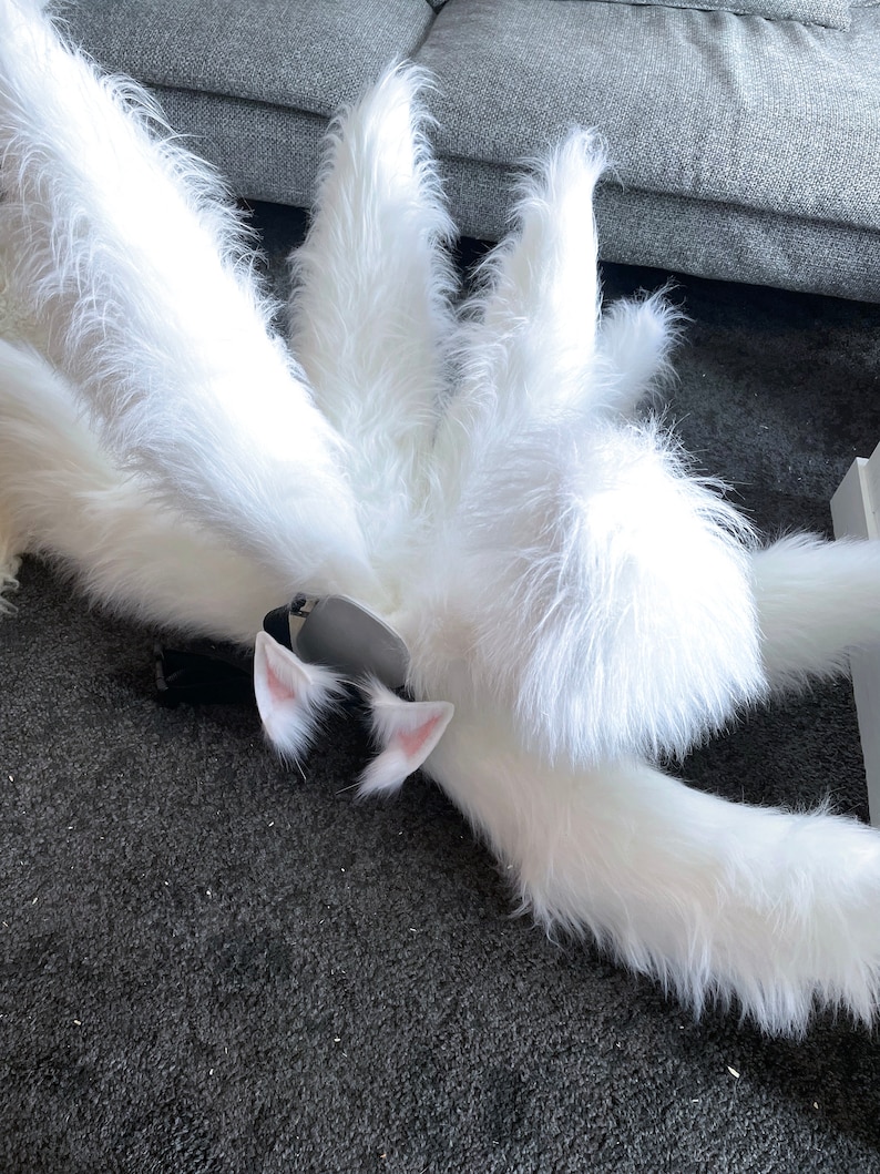 Op de afbeelding: Een pluizige witte vossenstaart met meerdere secties, gecombineerd met bijpassende witte en roze kattenoren en een zwarte hoofdband. De imitatiebont is lang en getextureerd, wat een grillige esthetiek cre&euml;ert. Perfect voor cosplay of kostuumfeesten.