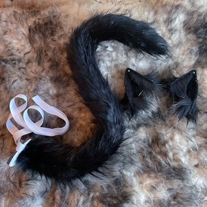 Könnte beinhalten: Ein schwarzes Kunstfell-Schwanz- und Ohren-Set mit weißem Band. Der Schwanz ist lang und gebogen, und die Ohren sind dreieckig mit einem schwarzen und grauen Farbverlauf. Das Set ist für Cosplay oder Kostüme gedacht.