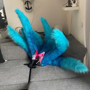 Op de afbeelding: Een blauwe en turquoise faux fur staart met meerdere pluizige secties. De staart is gecombineerd met roze en blauwe kattenoren met bloemaccenten en witte linten. Het kostuumaccessoire ligt op een grijze bank.