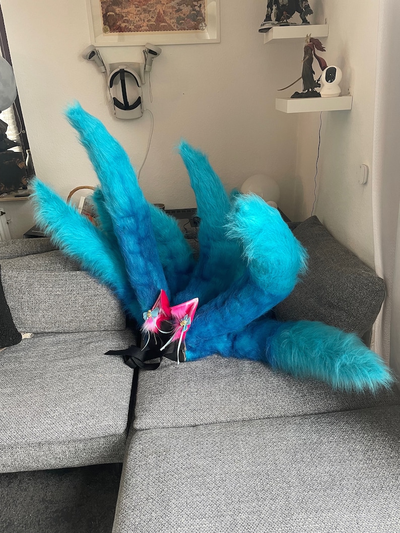 Op de afbeelding: Een blauwe en turquoise faux fur staart met meerdere pluizige secties. De staart is gecombineerd met roze en blauwe kattenoren met bloemaccenten en witte linten. Het kostuumaccessoire ligt op een grijze bank.