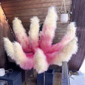 Op de afbeelding: Een pluizig rekwisiet met meerdere staarten in roze en cr&egrave;me. De nepbontstaarten zijn in een waaier gerangschikt, waarbij de roze kleur overgaat van de basis naar de punten. Het rekwisiet wordt op een metalen standaard gepresenteerd.