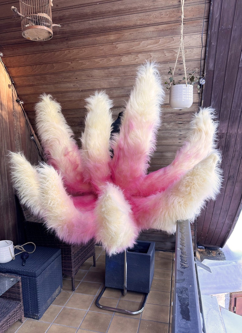 Op de afbeelding: Een pluizig rekwisiet met meerdere staarten in roze en cr&egrave;me. De nepbontstaarten zijn in een waaier gerangschikt, waarbij de roze kleur overgaat van de basis naar de punten. Het rekwisiet wordt op een metalen standaard gepresenteerd.