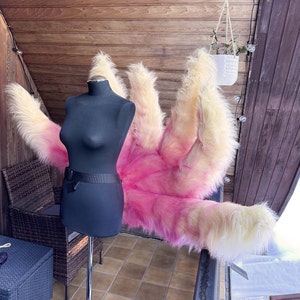 Op de afbeelding: Een zwarte paspop met een zwarte rok en riem staat naast een groot, pluizig kostuumstuk. Het kostuum is voornamelijk roze en geel, met een harige textuur. De achtergrond omvat een houten muur en een kleine hangplant.