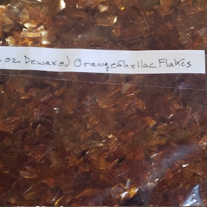 May include: A clear plastic bag filled with orange shellac flakes. A white label on the bag reads "2 oz. Dewaxed Orange Shellac Flakes". The flakes have a translucent, amber appearance.