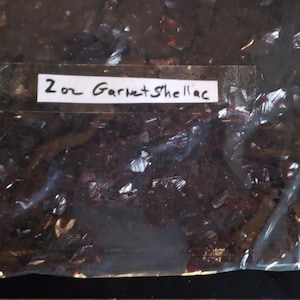 May include: A clear plastic bag filled with garnet shellac flakes. The bag has a white label with the handwritten text "2 oz Garnet Shellac". The shellac flakes are dark reddish-brown and appear shiny.