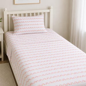 May include: White bed linens with a repeating pattern of pink and orange rainbows. The set includes a fitted sheet, flat sheet, and pillowcase. The bed frame is white, and the room has a neutral color scheme.