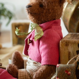 Pu&ograve; includere: Un orsacchiotto di peluche marrone che indossa una giacca di lino rosa con un papillon verde e pantaloni a quadri beige. L'orso &egrave; seduto su una superficie di juta marrone.