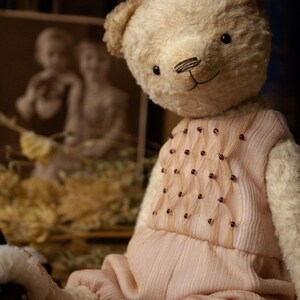 May include: A cream-colored teddy bear wearing a pink dress with a beaded design. The bear is sitting on a brown surface.