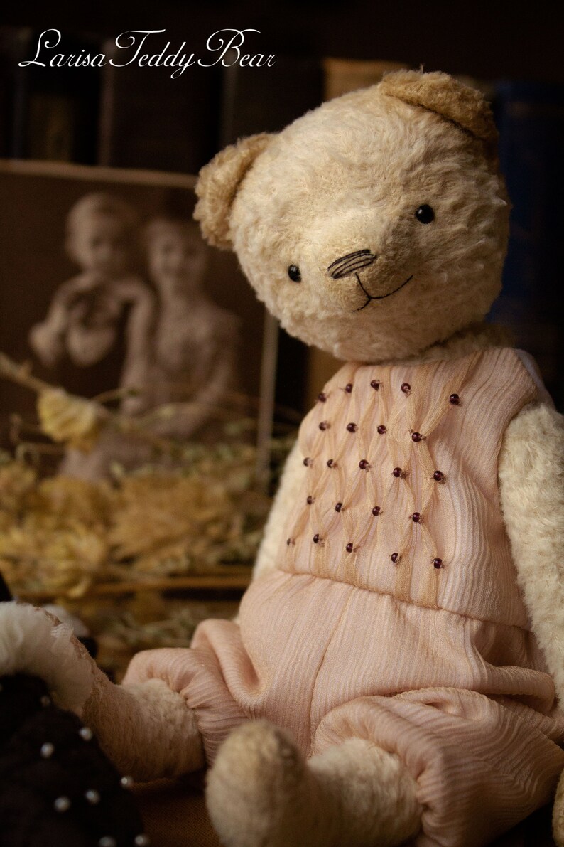 May include: A cream-colored teddy bear wearing a pink dress with a beaded design. The bear is sitting on a brown surface.